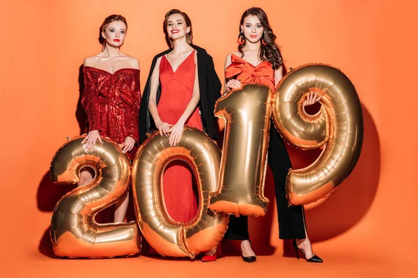 Ragazze attraenti sorridenti in abiti da festa alla moda che tengono palloncini 2019 su arancione, concetto di nuovo anno — Foto stock