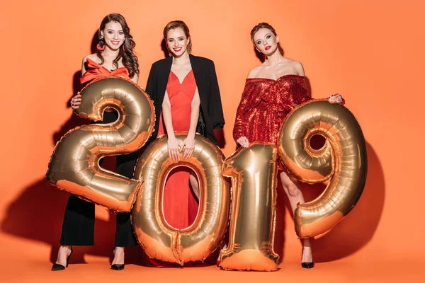 Счастливые красивые девушки в модных вечерних платьях с золотыми воздушными шарами 2019 года на оранжевой, новогодней концепции — стоковое фото