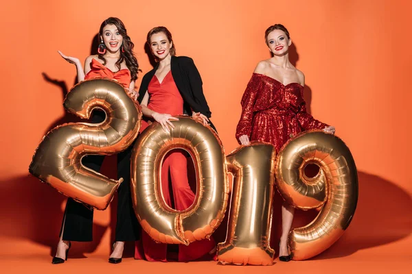 Surprised attractive girls in stylish party clothes holding 2019 balloons and looking at camera on orange — Stock Photo