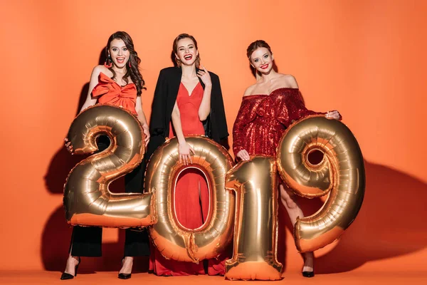 Улыбающиеся привлекательные девушки в стильной одежде для вечеринок с воздушными шарами 2019 года на апельсине — стоковое фото
