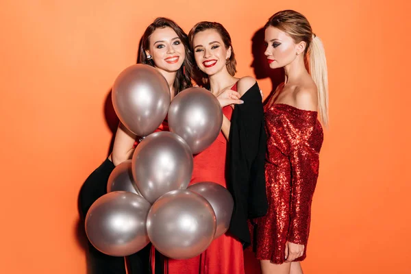 Smiling attractive girls in stylish party clothes holding bundle of grey balloons isolated on orange — Stock Photo