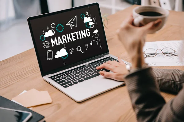 Cropped view of businesswoman with coffee cup typing on laptop with marketing icons — Stock Photo