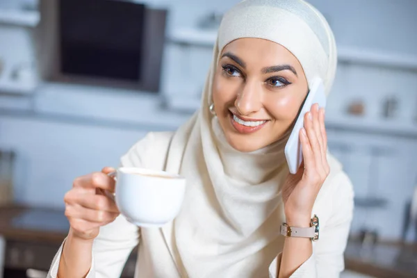 Mujer musulmana sonriente sosteniendo taza de café y hablando por teléfono inteligente - foto de stock