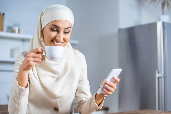 Улыбающаяся мусульманка держит чашку кофе и пользуется смартфоном дома — стоковое фото