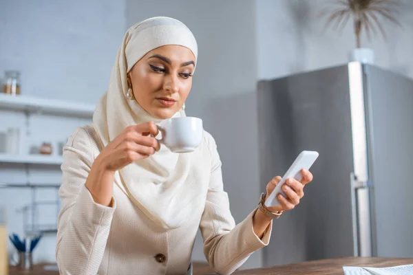 Mujer musulmana joven sosteniendo la taza de café y el uso de teléfono inteligente en casa - foto de stock