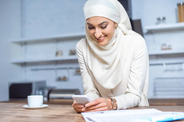 Hermosa mujer joven musulmana sonriente usando teléfono inteligente en casa - foto de stock