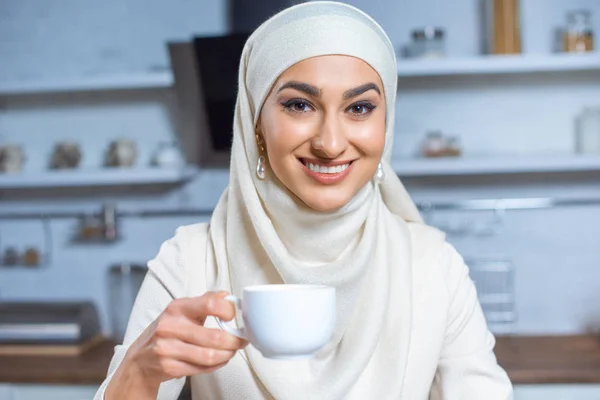 Hermosa feliz joven musulmana mujer sosteniendo la taza de café y sonriendo a la cámara - foto de stock