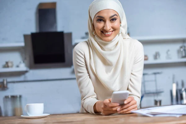 Hermosa joven musulmana mujer usando teléfono inteligente y sonriendo a la cámara - foto de stock