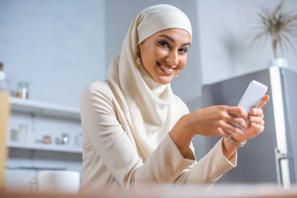 Молодая мусульманка, пользующаяся смартфоном и улыбающаяся в камеру — стоковое фото