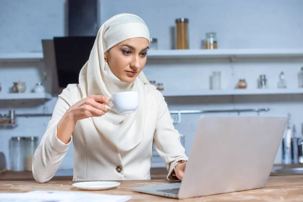 Молодая мусульманка с чашкой кофе и ноутбуком дома — стоковое фото