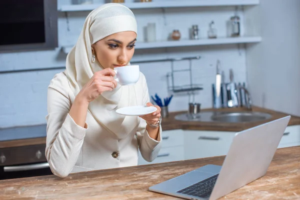 Молодая мусульманка пьет кофе и пользуется ноутбуком дома — стоковое фото