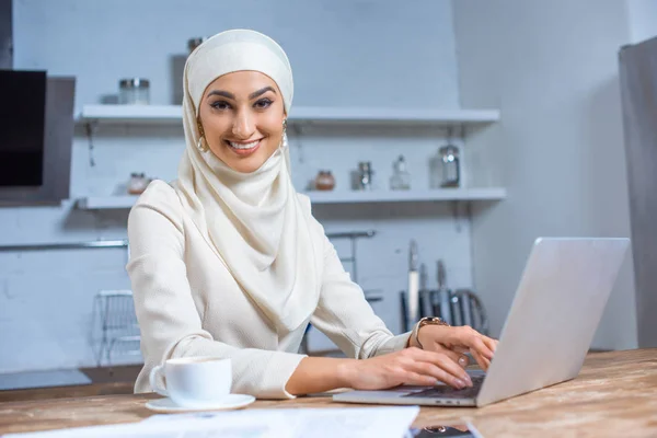 Bella giovane donna musulmana utilizzando il computer portatile e sorridente alla fotocamera — Foto stock