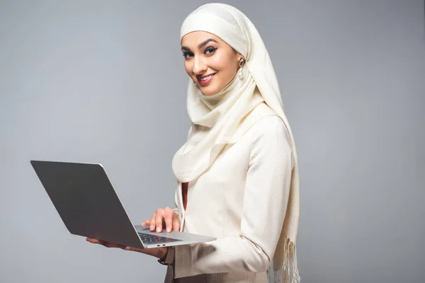 Young muslim woman using laptop and smiling at camera isolated on grey — Stock Photo