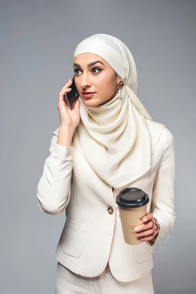 Jovem mulher muçulmana falando por smartphone e segurando copo de papel isolado em cinza — Fotografia de Stock
