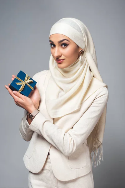 Молода мусульманка тримає подарунок і посміхається на камеру ізольовано на сірому — стокове фото