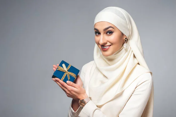 Happy young young muslim woman holding present and smiling at camera isolated on grey — Stock Photo