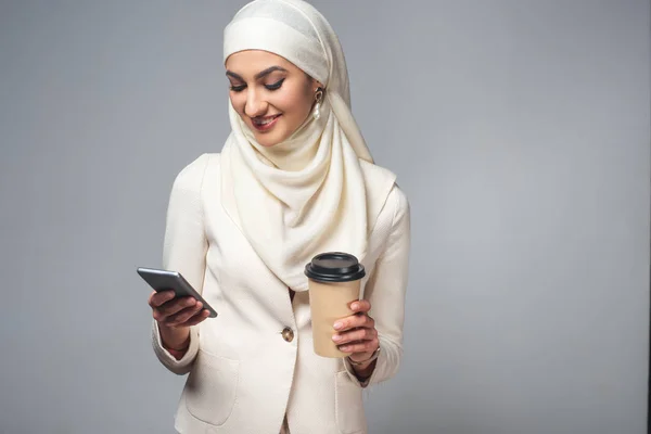 Sorridente giovane donna musulmana in possesso di caffè per andare e utilizzando smartphone isolato su grigio — Foto stock