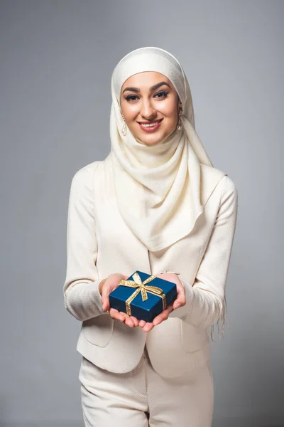 Felice giovane donna musulmana in possesso di una scatola regalo e sorridente alla fotocamera isolata sul grigio — Foto stock