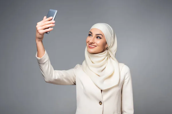 Красивая улыбающаяся мусульманка делает селфи со смартфоном, изолированным на сером — стоковое фото