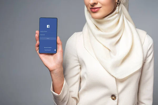 Recortado disparo de mujer musulmana sonriente sosteniendo teléfono inteligente con aplicación de facebook aislado en gris - foto de stock