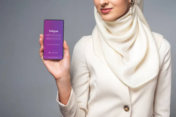 Recortado disparo de mujer musulmana sonriente sosteniendo teléfono inteligente con aplicación instagram aislado en gris - foto de stock