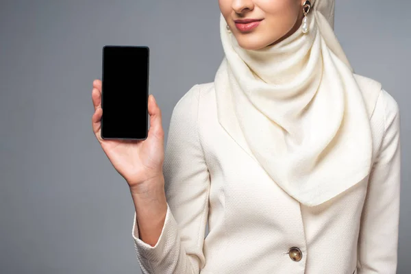 Abgeschnittene Aufnahme einer lächelnden muslimischen Frau mit Smartphone und leerem Bildschirm isoliert auf grau — Stockfoto