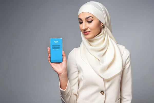 Hermosa sonriente joven musulmana sosteniendo teléfono inteligente con aplicación skype aislado en gris - foto de stock