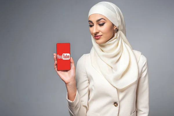 Beautiful smiling young muslim woman holding smartphone with youtube app isolated on grey — Stock Photo