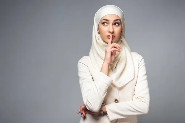 Молодая мусульманка, стоящая с пальцем на губах и глядя в сторону, изолированная на сером — стоковое фото