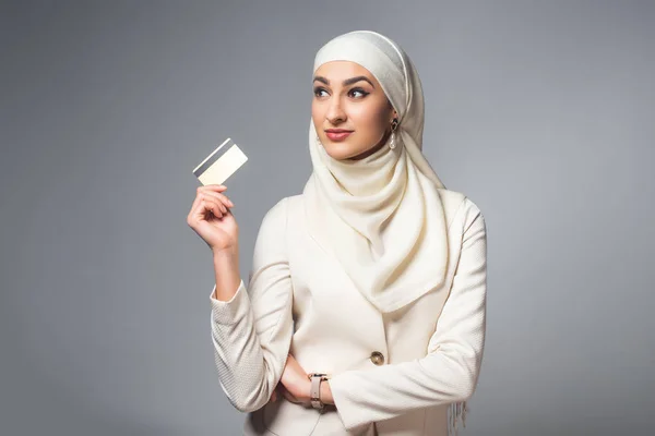 Hermosa mujer musulmana sonriente sosteniendo tarjeta de crédito y mirando hacia otro lado aislado en gris - foto de stock