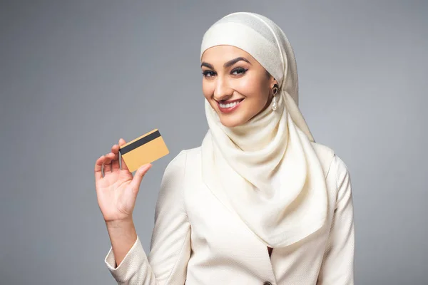 Молодая мусульманка, держащая кредитку и улыбающаяся в камеру, изолированную на серой — стоковое фото