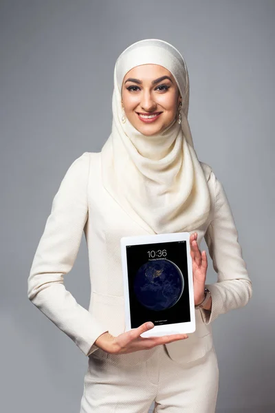 Joven mujer musulmana sosteniendo la tableta digital y sonriendo a la cámara aislada en gris — Stock Photo