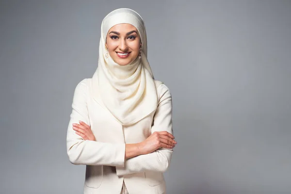 Portrait of beautiful young muslim woman standing with crossed arms and smiling at camera isolated on grey — Stock Photo