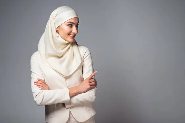 Sorridente giovane donna musulmana in piedi con le braccia incrociate e distogliendo lo sguardo isolato sul grigio — Foto stock