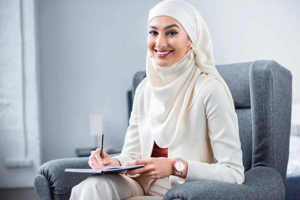 Красива молода мусульманка, що пише в блокноті і посміхається на камеру — стокове фото