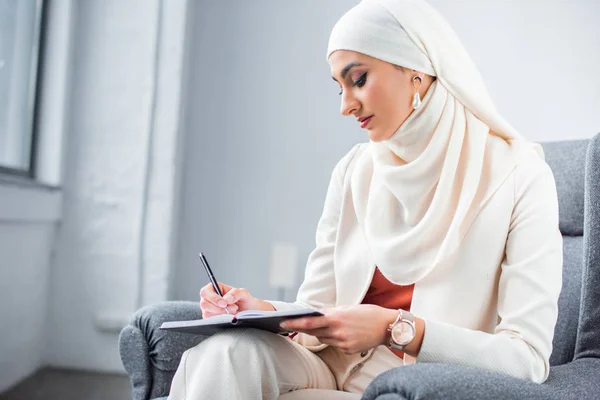 Schöne junge muslimische Frau sitzt im Stuhl und schreibt in Notizbuch — Stockfoto