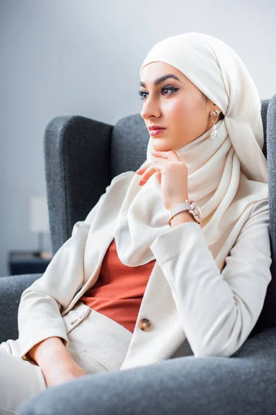 Hermosa mujer musulmana joven pensativo sentado en la silla y mirando hacia otro lado - foto de stock