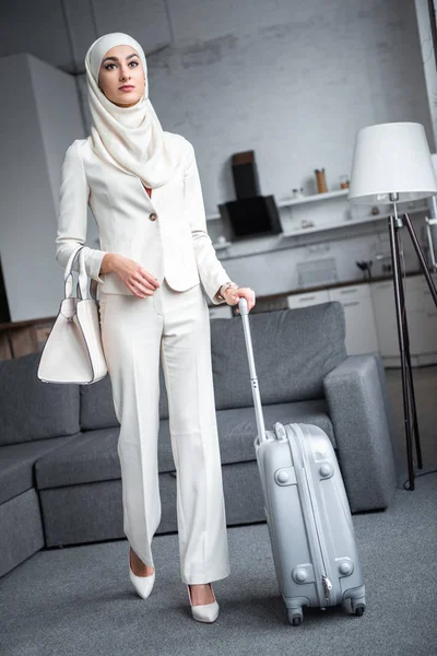 Jovem mulher muçulmana em terno branco segurando mala e olhando para longe em casa — Fotografia de Stock
