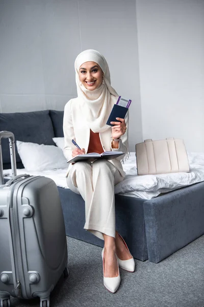 Belle jeune femme musulmane tenant passeport avec carte d'embarquement et souriant à la caméra à la maison — Photo de stock
