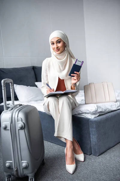 Bela mulher muçulmana segurando passaporte com cartão de embarque e sorrindo para a câmera no quarto — Fotografia de Stock
