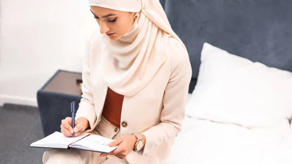 High angle view of young muslim woman sitting on bed and writing in notebook — Stock Photo