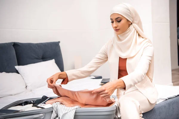 Jeune femme musulmane assise sur le lit et emballant des vêtements dans la valise — Photo de stock