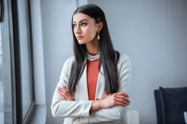 Schönes elegantes Mädchen, das mit verschränkten Armen steht und zum Fenster schaut — Stockfoto