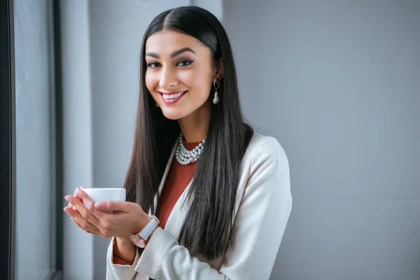 Belle jeune femme tenant tasse de café et souriant à la caméra — Photo de stock