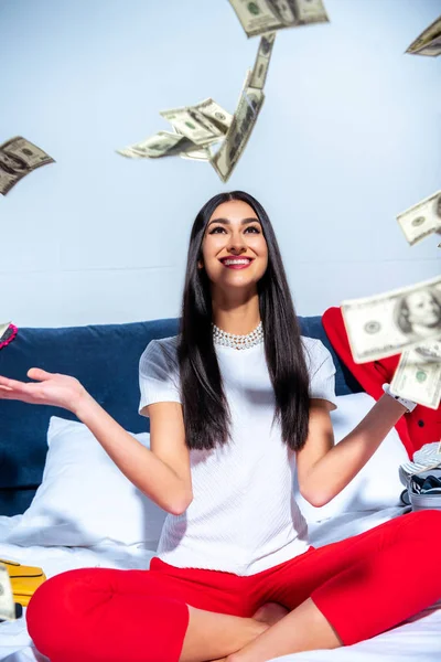 Fröhliche junge brünette Frau sitzt auf dem Bett und wirft Dollarnoten — Stockfoto