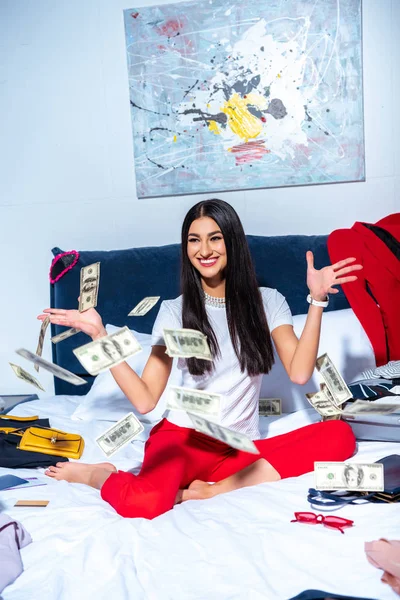 Beautiful happy young woman sitting on bed and throwing dollar banknotes — Stock Photo