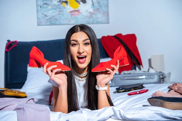Excited young woman holding stylish red shoes and smiling at camera while lying on bed before tip — Stock Photo