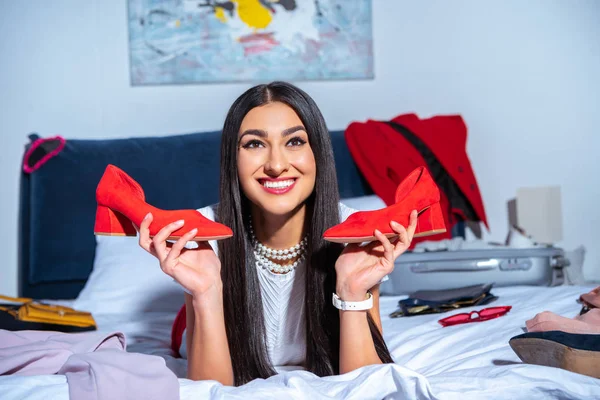 Красива усміхнена молода жінка тримає модне червоне взуття, лежачи на ліжку перед поїздкою — стокове фото