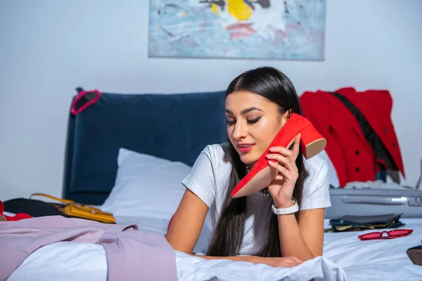 Beautiful smiling young woman holding red shoe and lying on bed before trip — Stock Photo