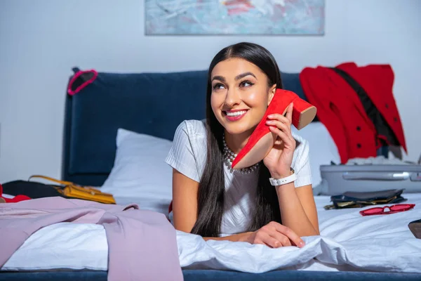 Hermosa chica sonriente sosteniendo zapato rojo y acostado en la cama antes del viaje - foto de stock